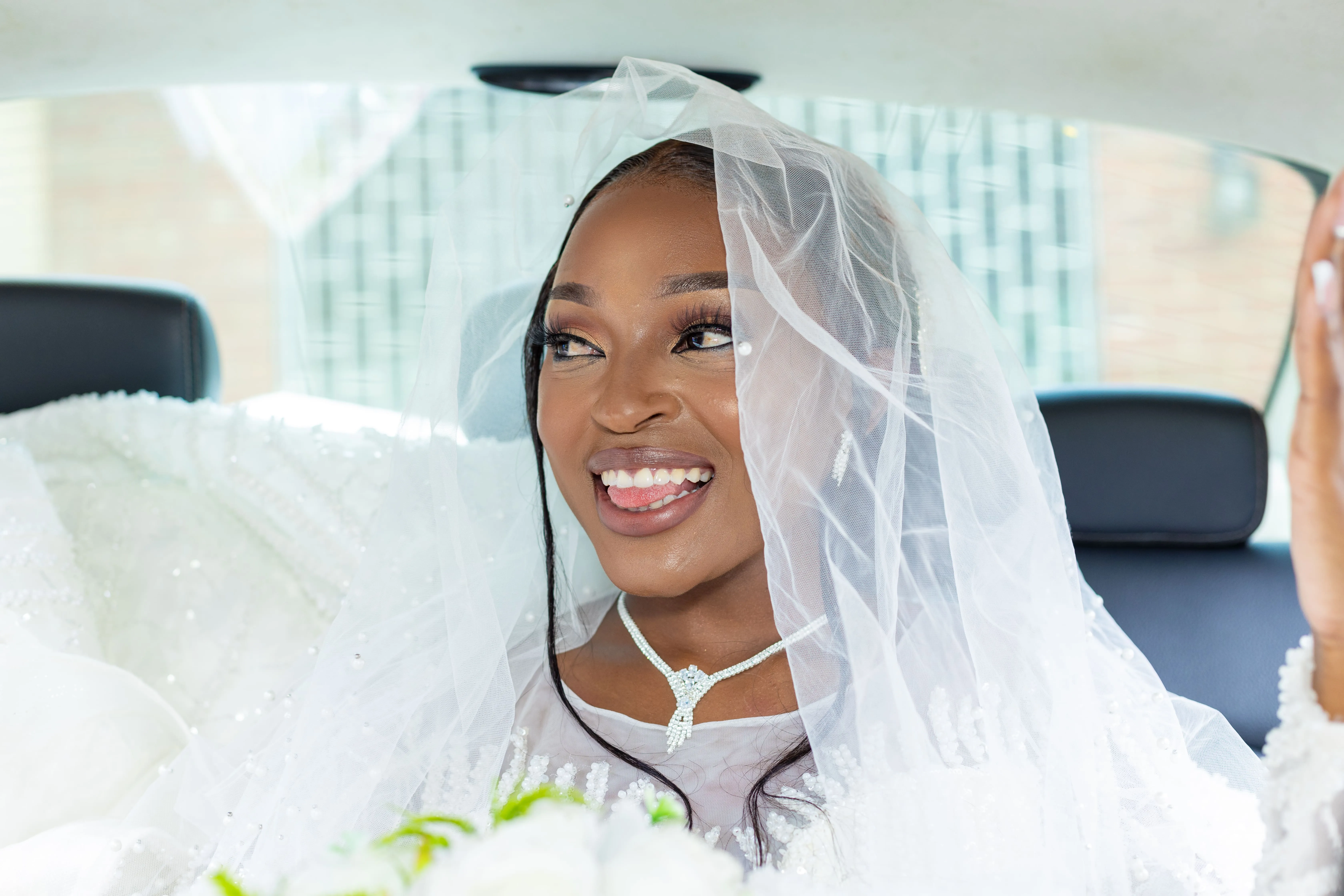 Smiling bride wearing a pearl-embellished veil and necklace sitting in the backseat of a car.