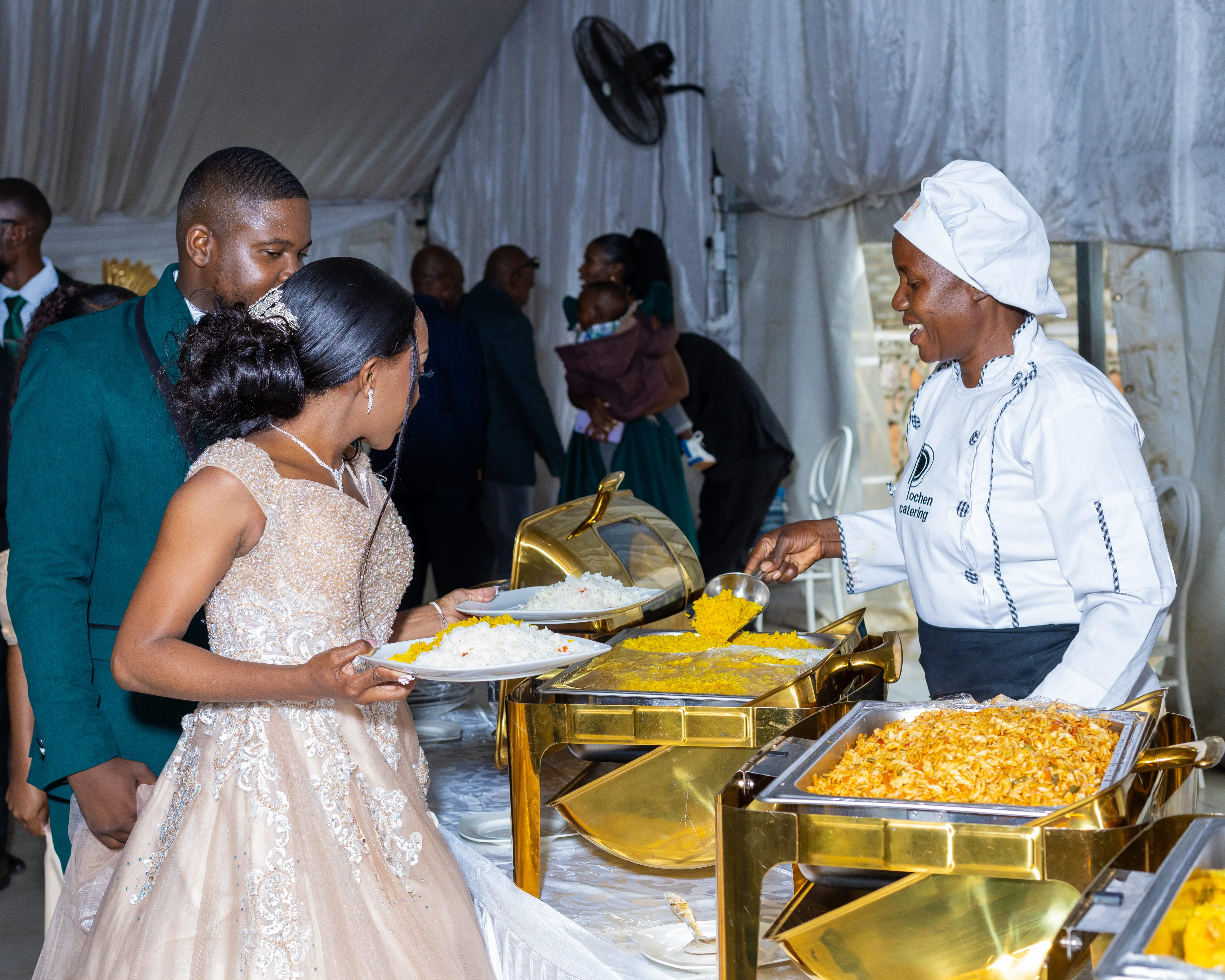 Bride and groom serving themselves food from a buffet with a smiling chef scooping rice in the background.