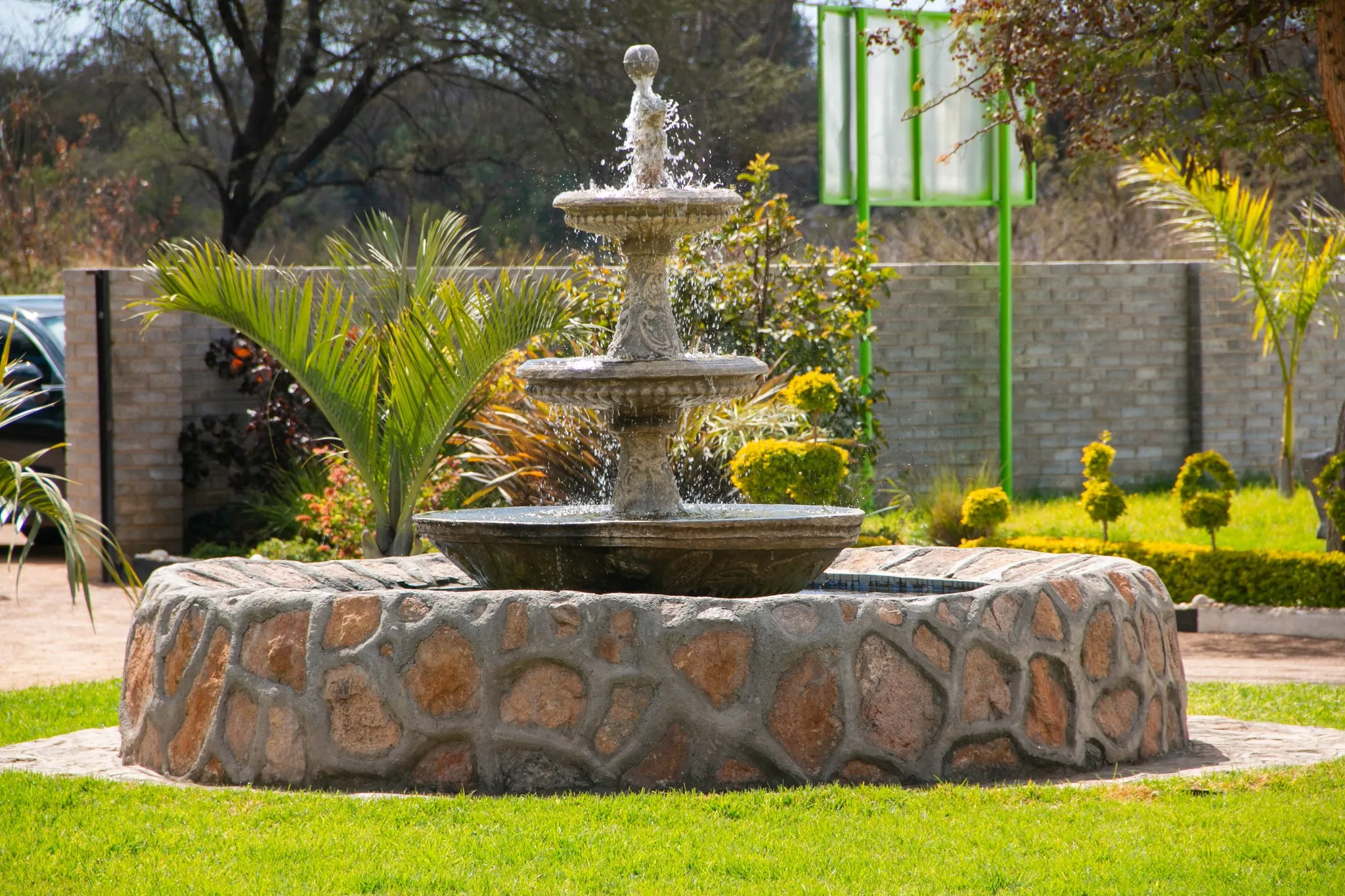 Three-tier stone water fountain with water splashing, surrounded by green grass and tropical plants in a garden.