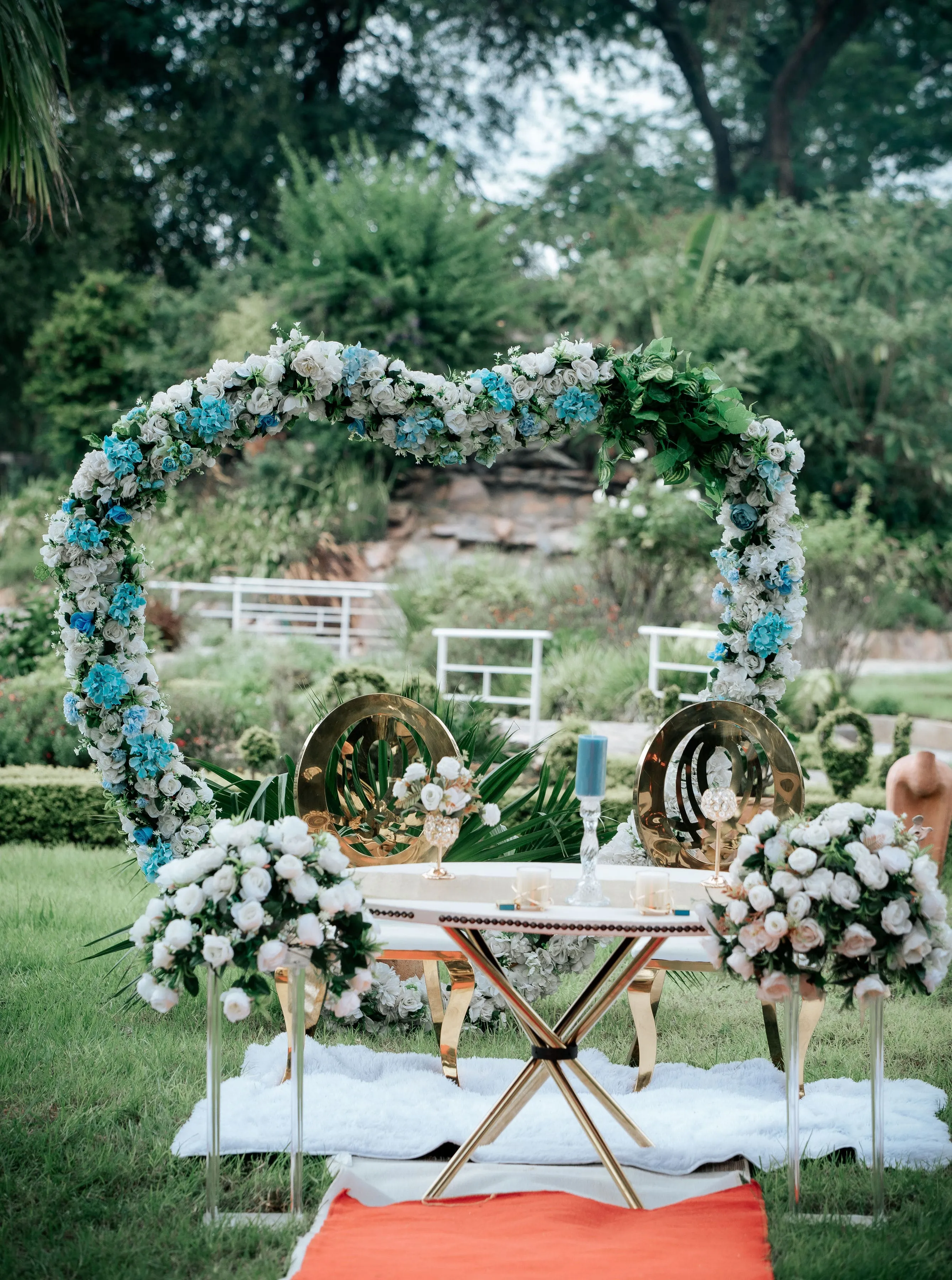 Outdoor wedding setup with a heart-shaped floral arch of white and blue flowers, two golden chairs, a white table with decorations, and floral arrangements on grass.