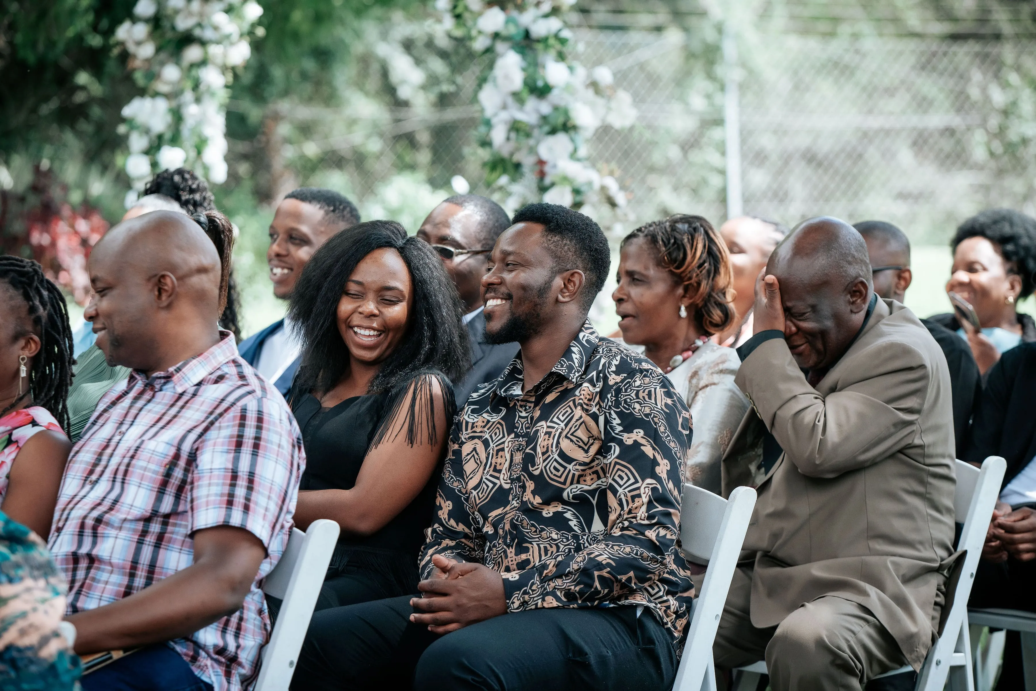 Happy group of well-dressed people smiling and laughing while seated outdoors on white chairs.