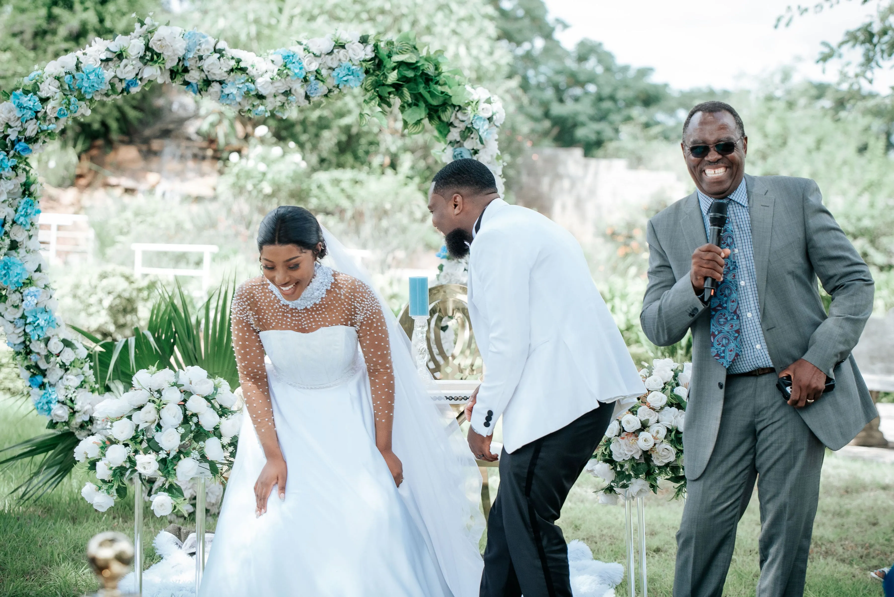 Bride and groom laughing during an outdoor wedding ceremony with an officiant holding a microphone.