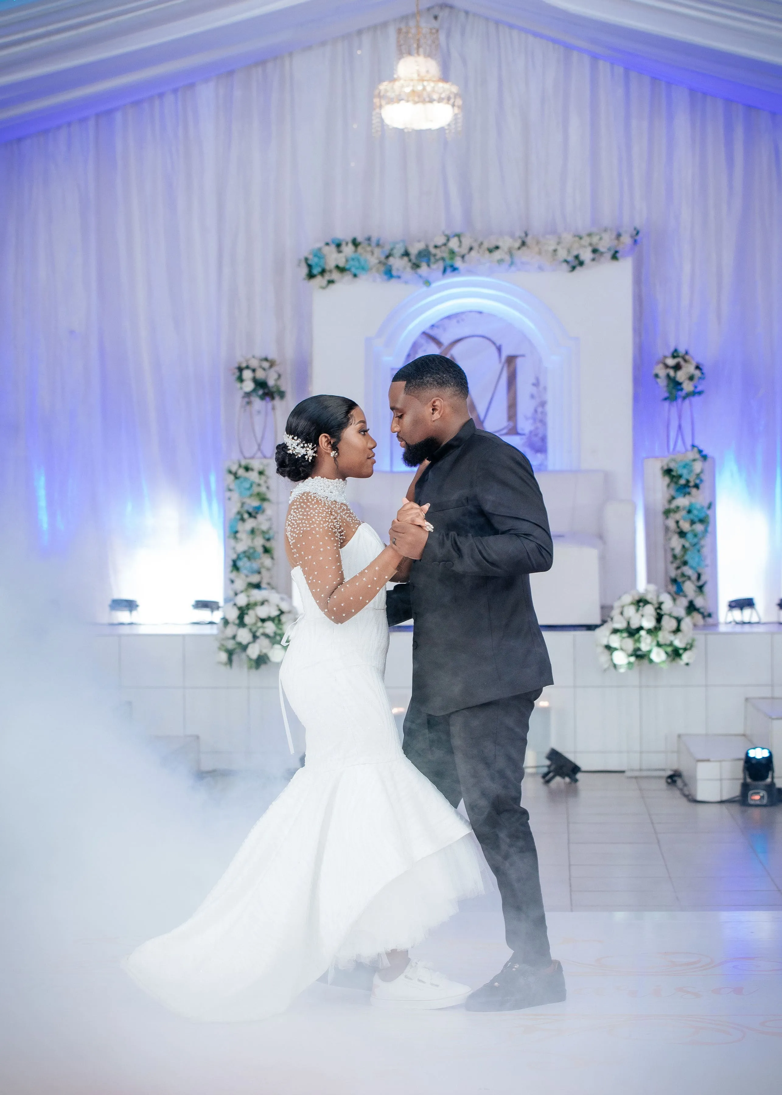 Bride in a white dress and sneakers dancing with groom in black suit at a wedding with floral decorations and blue lighting.