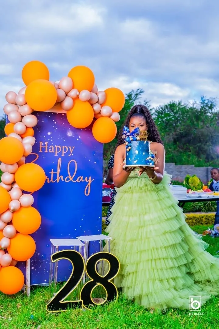 Woman in green tiered dress holding a blue birthday cake with decorations, standing next to a birthday sign with orange and beige balloons and a large illuminated '28' number on grass.